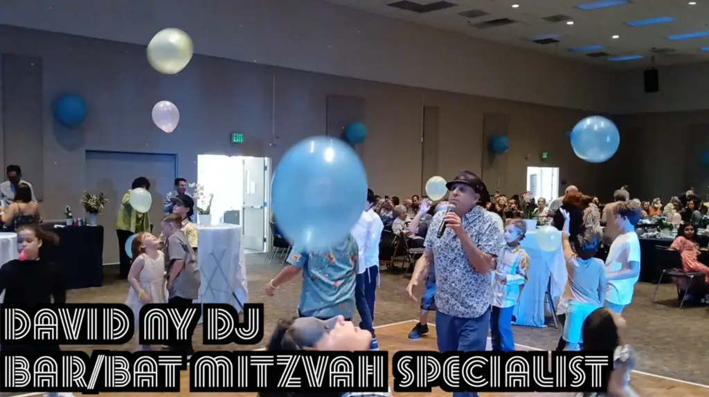 A crowded event venue with people of various ages celebrating an occasion. The room is decorated with blue and white balloons, creating a festive atmosphere. People are seen enjoying themselves, some holding or interacting with the balloons.